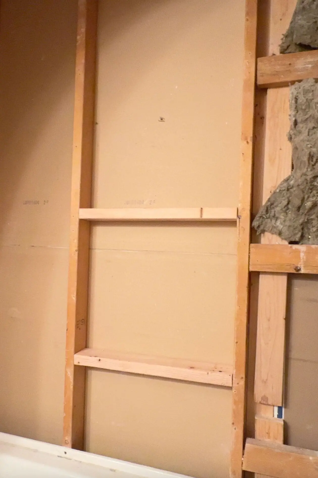 An unfinished bathroom wall with exposed wooden studs and insulation on one side. The studs include a horizontal support frame, for creating a recessed shower niche or additional structural stability during the renovation process.